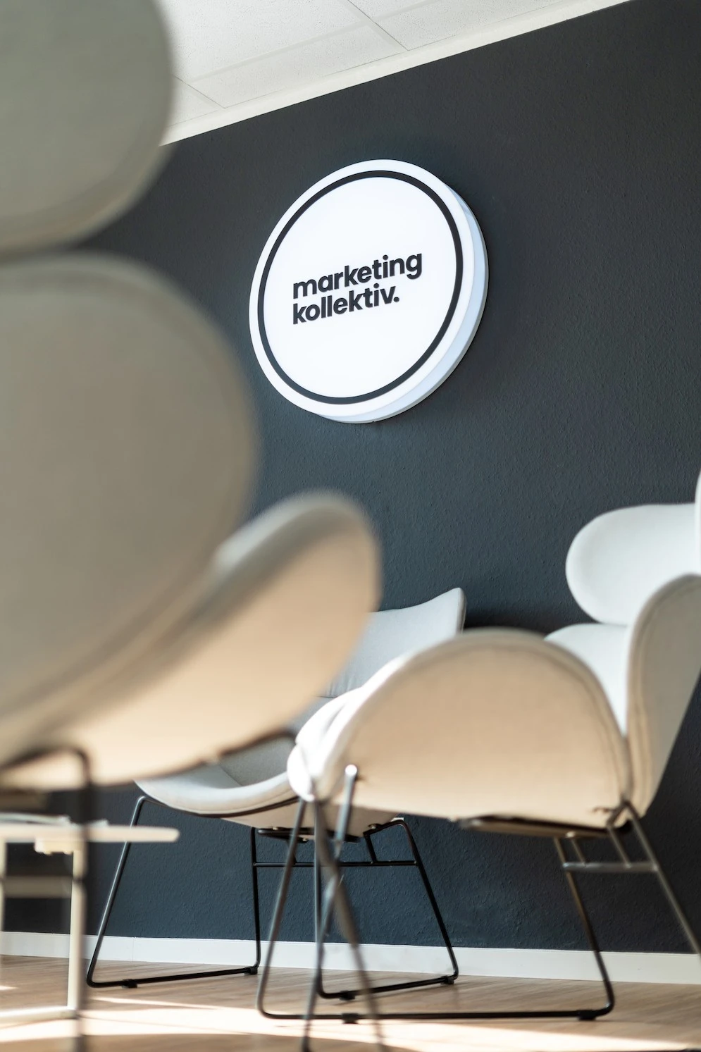 Runde Leuchttafel mit dem Logo von marketingkollektiv an dunkler Wand, davor helle Sessel in der Lounge